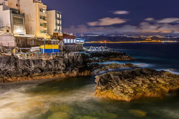 Water washing around coastal rocks beneath buildings at dusk.