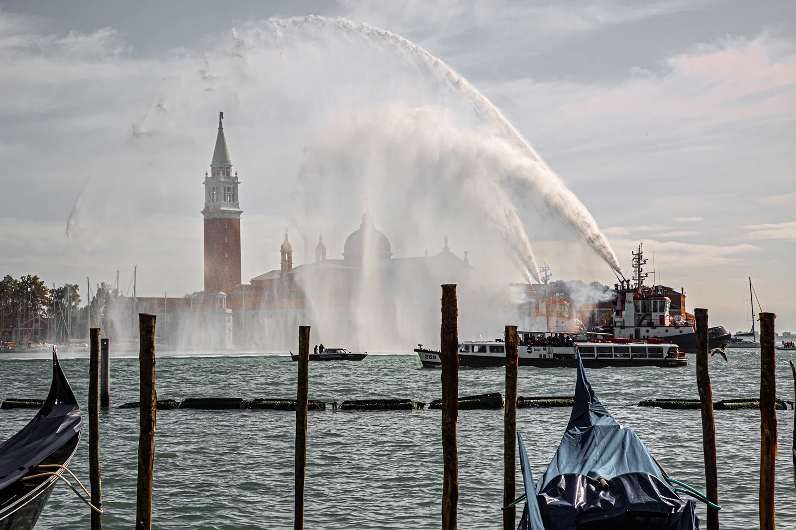 Boats spray arcs of water across a waterfront, with a bell tower in the distance.
