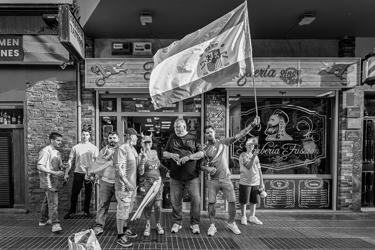 A group of football supporters holding a Spanish flag outside a bar, photographed in black and white.