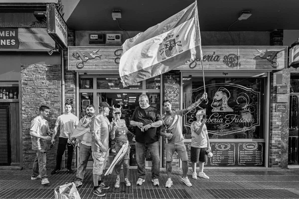 A group of football supporters holding a Spanish flag outside a bar, photographed in black and white.