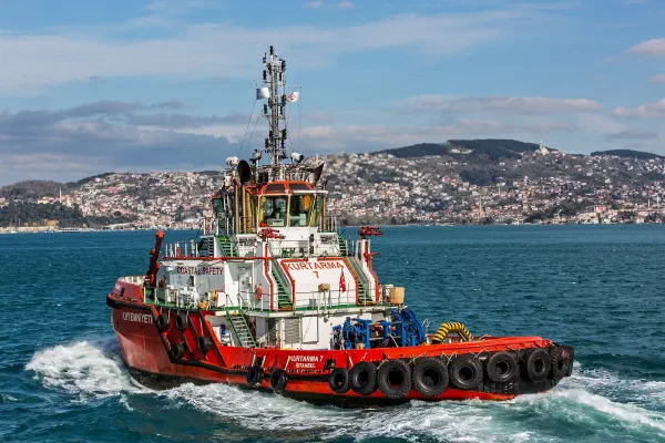 A working tugboat crossing blue water with a coastal city behind it.