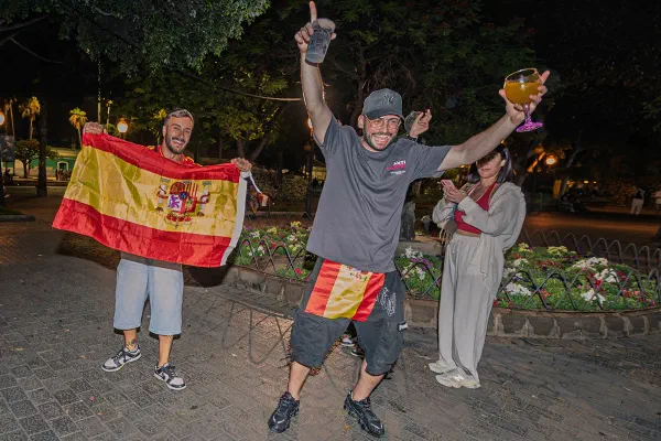 Three supporters holding flags and raising drinks outdoors at night.